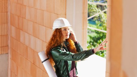 Woman with hard hat and blueprint-casing inside a building shell touching a brick
